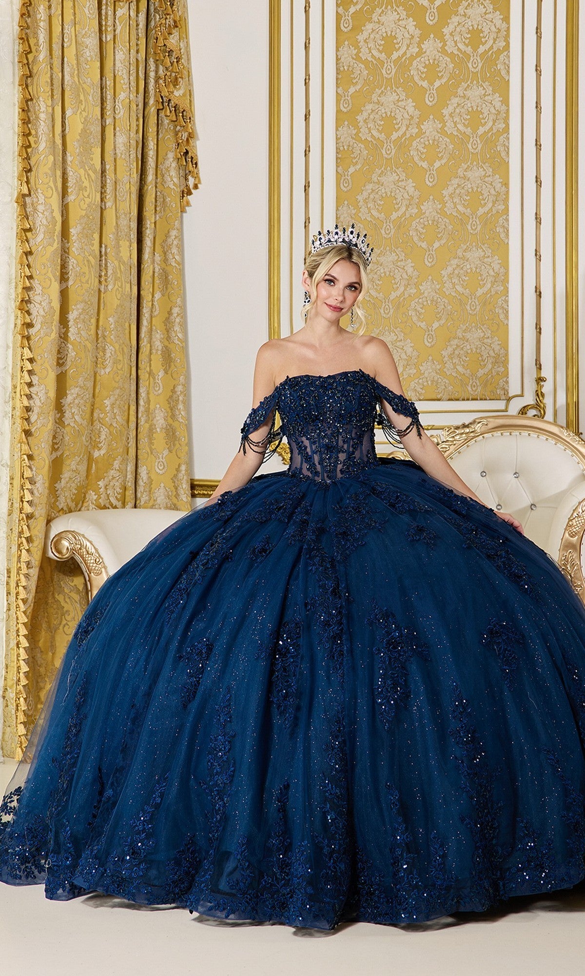 A woman in the Dancing Queen Quinceanera Dress 5141 in navy, featuring sequin and lace details, sits elegantly on a cream sofa in an ornate, gold-accented room, her look completed with a tiara.